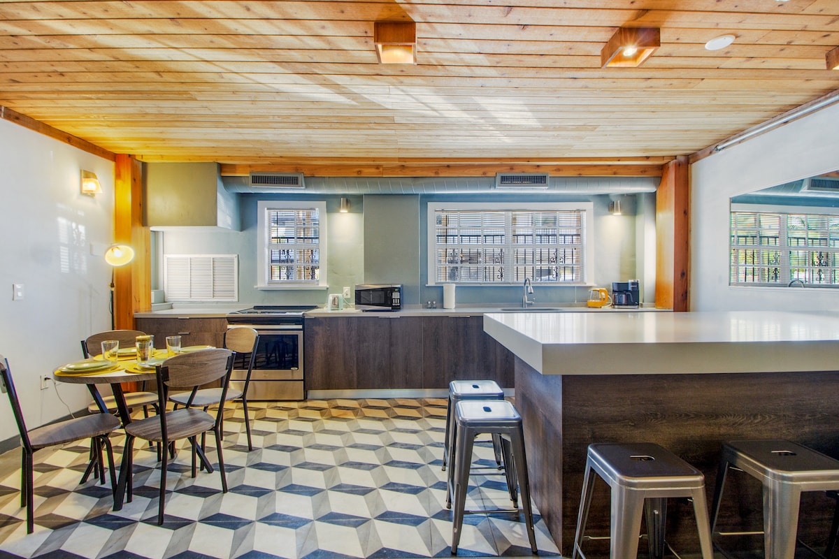 A modern kitchen area features a gray island with bar stools and a circular dining table set for four. Cabinets with open shelving display dishware, while a microwave and oven are neatly integrated. A patterned tiled floor complements the warm wooden ceiling.