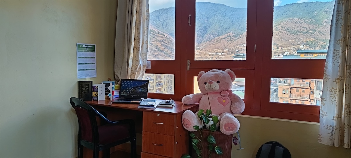 A workspace is situated next to large windows, offering views of the mountains. A desk holds a computer and stationery. A plush teddy bear rests on a chair and a potted plant adds a touch of greenery, enhancing the inviting atmosphere of the area.