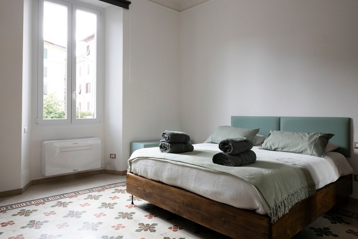 A spacious bedroom features a king-size bed topped with neatly arranged towels. Light walls reflect natural light from large windows, enhancing the serene ambiance. An air conditioning unit is positioned on the wall, while decorative flooring adds subtle character to the room.