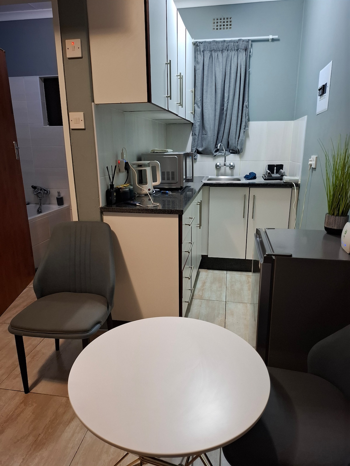 A compact kitchen area is showcased, featuring white cabinetry and a dark countertop. A small round table is positioned between two gray chairs. Natural light enters through a nearby window, and a microwave and kettle are visible on the counter.