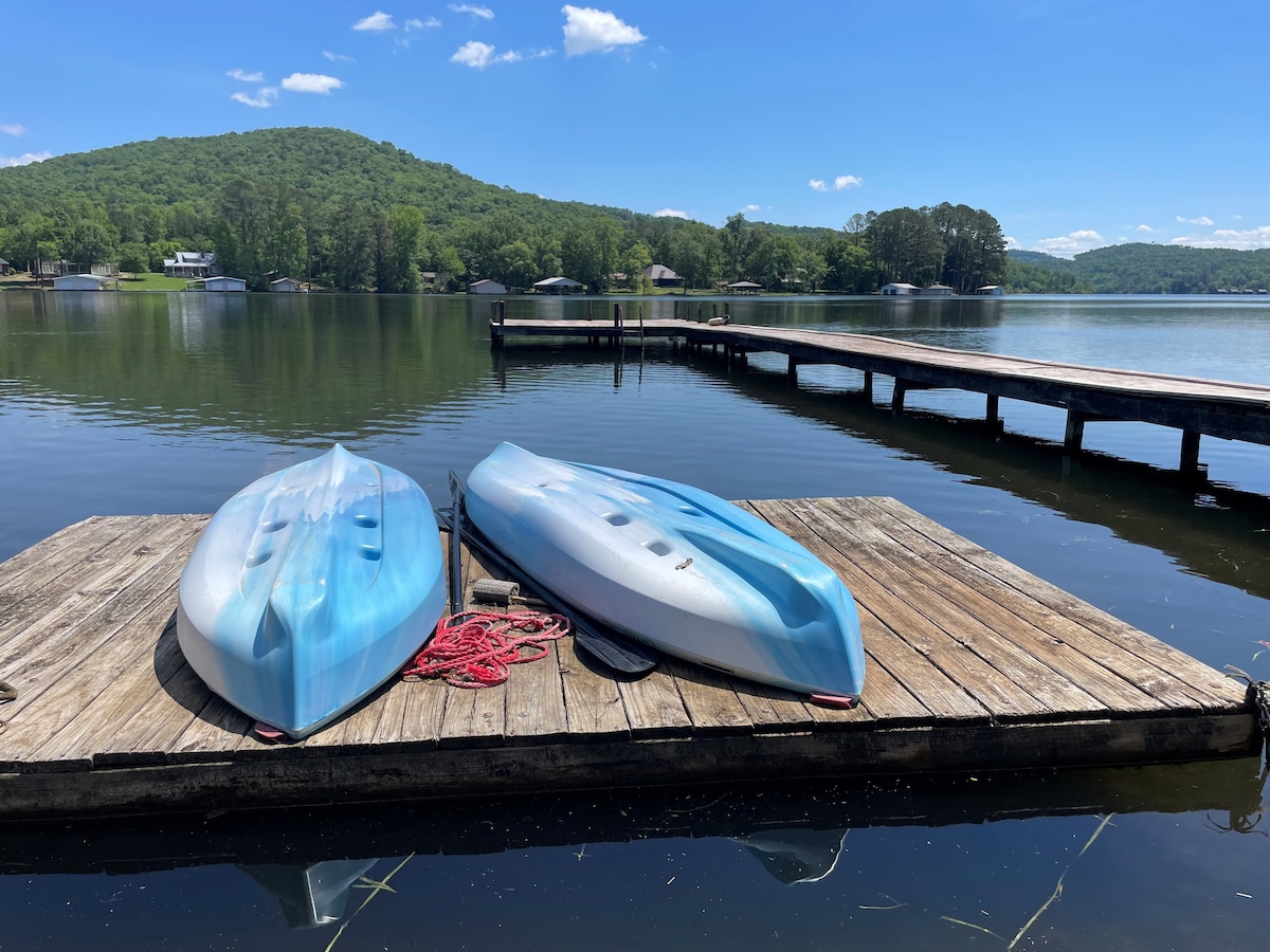 Two kayaks are positioned on a wooden dock beside a calm lake. The surrounding landscape features rolling green hills under a clear blue sky. The dock extends into the water, creating a serene atmosphere for outdoor activities.