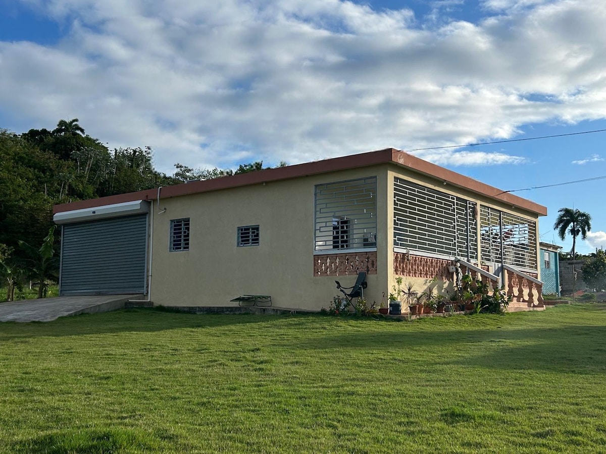 The exterior of a single-story building is shown, featuring a covered porch with decorative railing. Large windows offer ample natural light. A garage with a metallic door is visible on the left. Lush green grass surrounds the structure, enhancing the overall landscape.