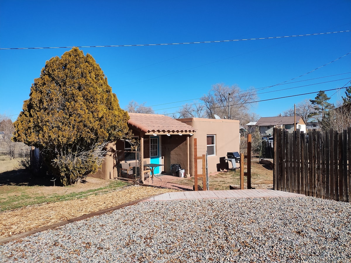 The exterior of the adobe-style house is shown, featuring a tiled roof and a welcoming porch area. A large tree provides shade, while a gravel path leads to the front entrance. A fenced yard encloses the space, with clear blue skies in the background.