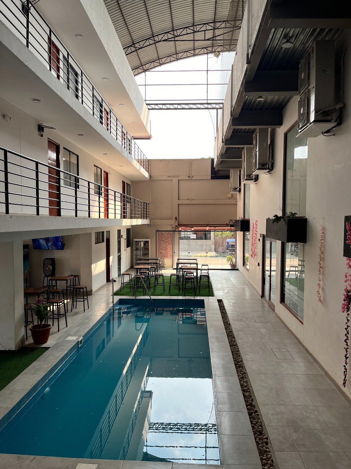 An open-air corridor features a clear swimming pool surrounded by lounge chairs and tables. The two-story structure showcases a modern design with large windows and sliding doors, allowing natural light to fill the space. Greenery is present, adding a touch of color to the surroundings.