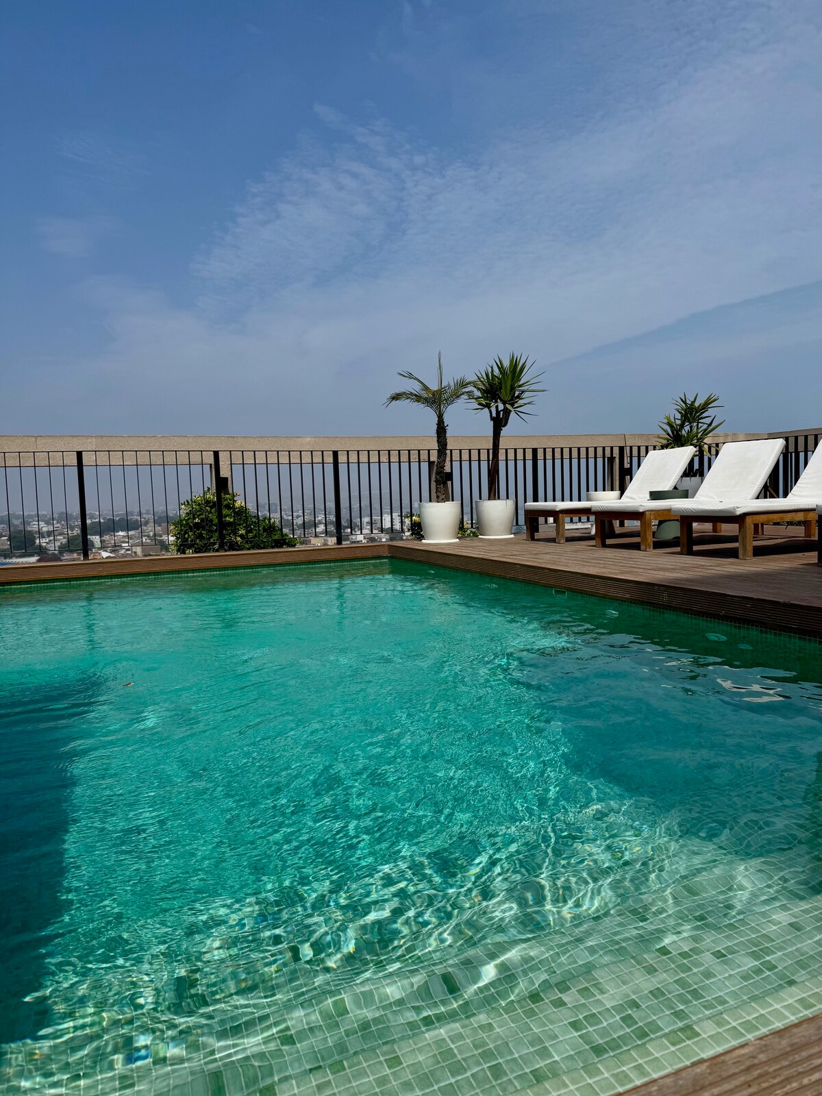 A rooftop pool is captured under a bright blue sky, showcasing clear turquoise water. Two lounge chairs are positioned beside the pool, along with potted plants adding greenery to the space. The horizon displays distant city views, enhancing the relaxing atmosphere.