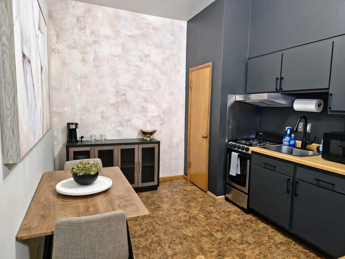 A modern kitchen area features gray walls and a warm wood dining table with a small green centerpiece. A dark wooden cabinet stands alongside the table. The kitchen is equipped with dark cabinets, a stovetop, and a microwave, offering a functional and stylish cooking space.