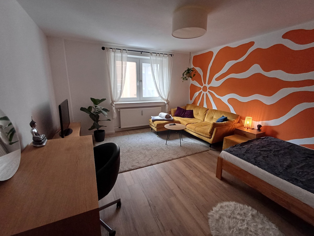 A vibrant living area features an orange accent wall with a swirling design. A spacious yellow couch is paired with a round coffee table, accompanied by a plant. A light-toned area rug sits on the wooden floor, and a window allows natural light to fill the room.