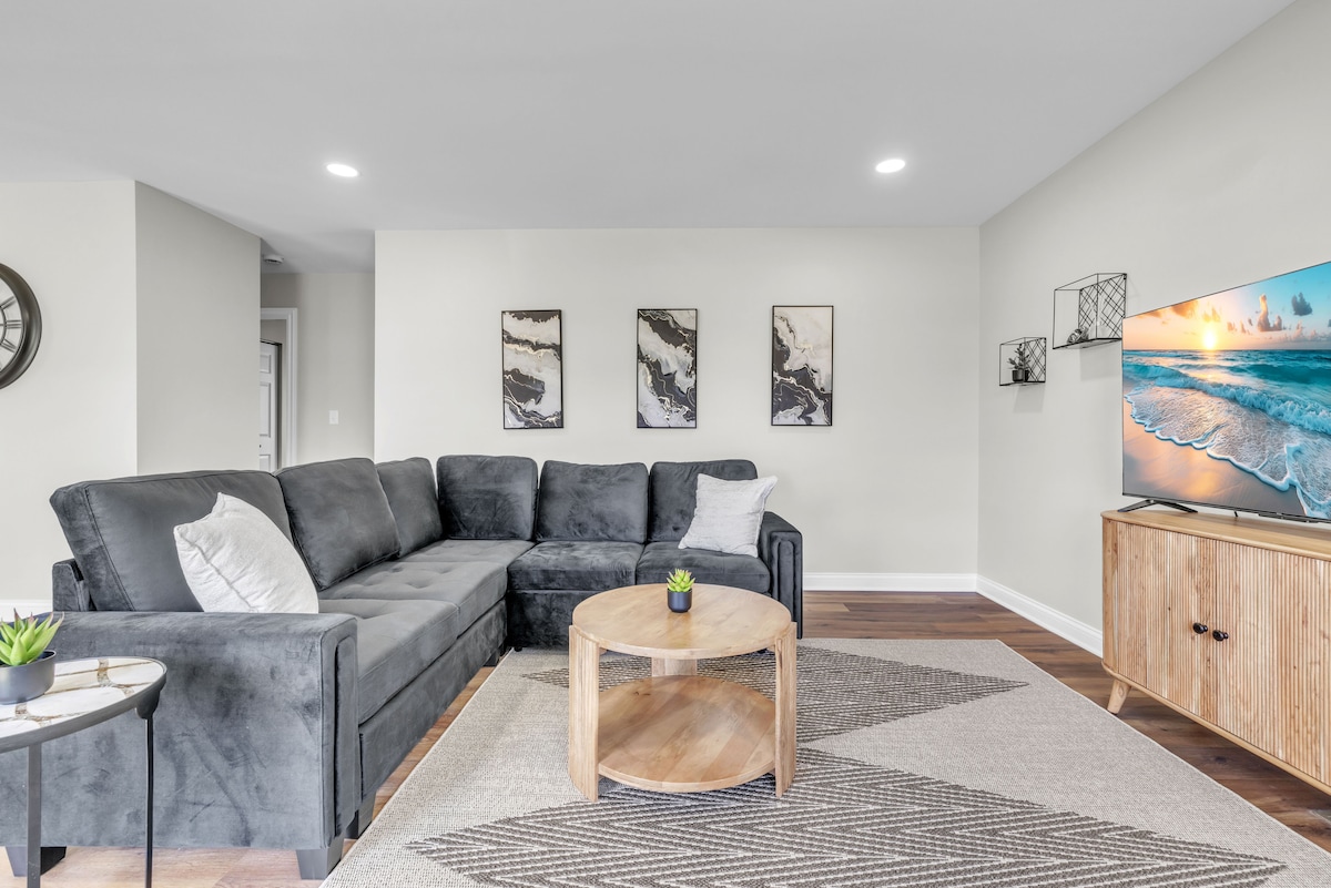 The living room features a spacious, gray sectional sofa adorned with decorative pillows. A round coffee table sits at the center, accompanied by a small side table with a potted plant. A 55” TV is positioned on a wooden cabinet, and framed artwork decorates the walls.