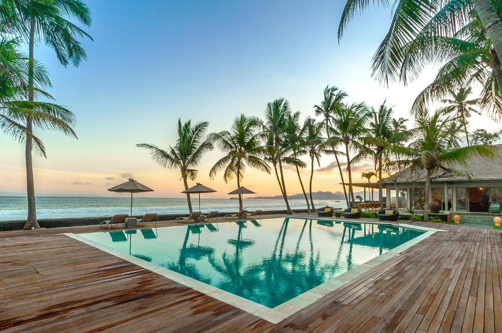 A tranquil outdoor area features a large swimming pool surrounded by tropical palm trees. Sun loungers and umbrellas are positioned near the pool's edge, offering shaded relaxation. In the background, the ocean is visible, enhancing the serene coastal setting during sunset.