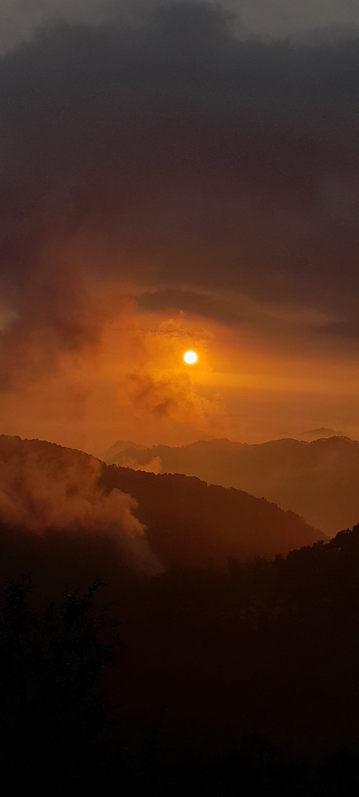 A serene sunset landscape displays rolling mountains shrouded in soft clouds. The sun casts a warm, golden hue over the scene, creating a peaceful and tranquil atmosphere as the day transitions into evening.