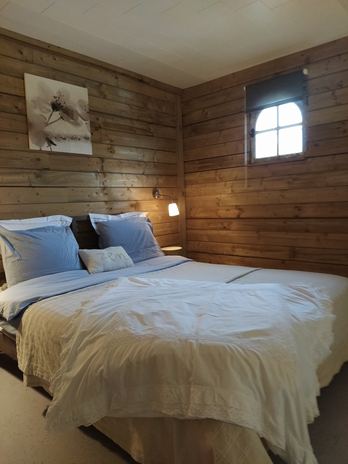 A wooden room features a spacious bed adorned with soft white linens and blue accent pillows. A bedside light provides gentle illumination. A small window allows natural light to enter, contributing to the cozy atmosphere of the space.