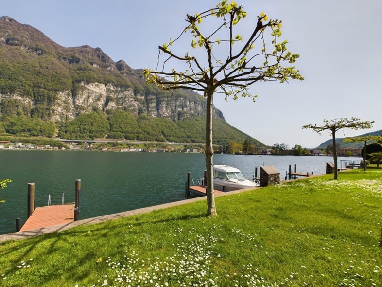 A serene lakeside view is presented, featuring a gentle slope of green grass leading to the water's edge. A small dock extends into the lake, and a boat is moored nearby. Rolling hills rise in the background, partially shaded by a tree.