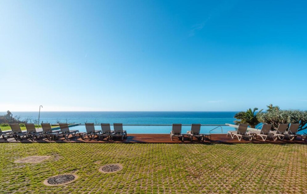 A tranquil outdoor space is presented, featuring numerous lounge chairs arranged along a wooden deck. The infinity pool extends towards the ocean, with a clear blue sky reflected in the water. Lush greenery is visible in the background.