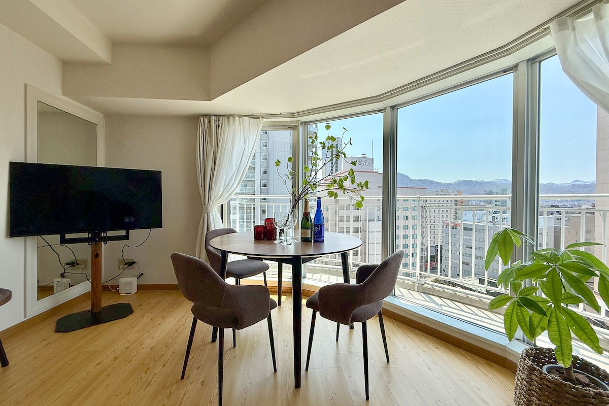 A bright dining area features a round table accompanied by four chairs, with a large window providing expansive views of the cityscape. Natural light floods the room, enhancing the sense of space. A television is mounted on the wall, and a potted plant adds a touch of greenery.