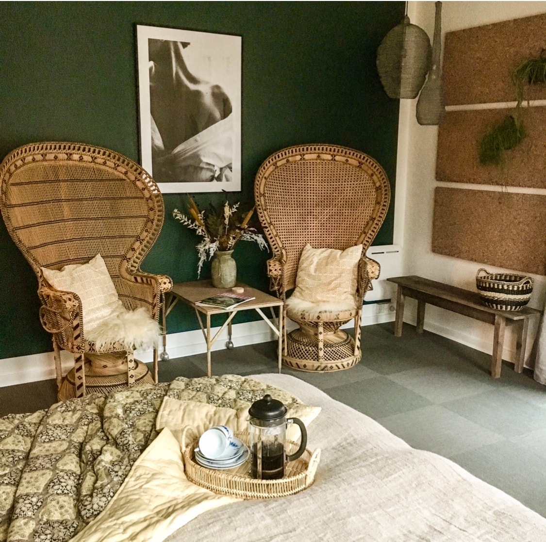 Two intricately woven chairs are positioned beside a wooden table, which holds a tea set and a French press. A soft, patterned blanket covers a spacious bed. A dark green wall serves as a backdrop to a large framed photograph, contributing to the calm ambiance.