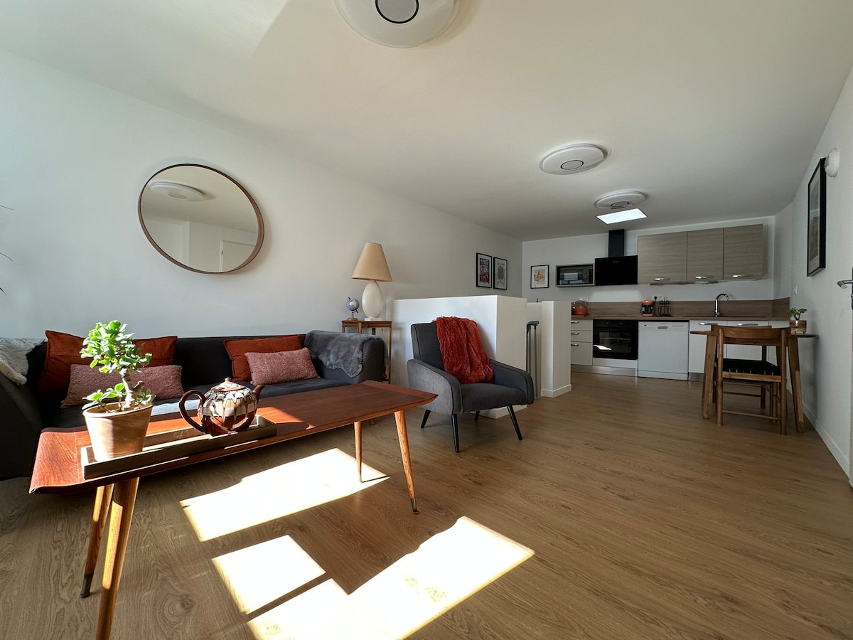 A cozy living area features a neutral color palette with a large mirror reflecting natural light. A coffee table sits prominently in front of a comfortable seating arrangement. An open kitchen and dining area are visible, showcasing modern appliances and a dining table with chairs.