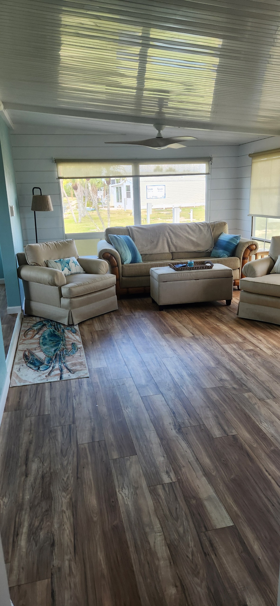 A light-filled living area features two upholstered chairs and a sofa adorned with decorative pillows. A ceiling fan is positioned above, while large windows offer a view of the surroundings. A decorative rug adds warmth to the wood-like floor.