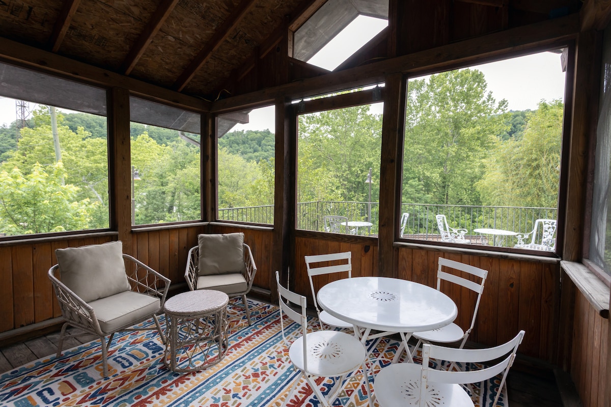 A screened-in porch features a round table surrounded by four white chairs. Two beige armchairs are positioned nearby on a patterned rug. Large windows showcase the surrounding greenery and distant hills, allowing natural light to brighten the space.