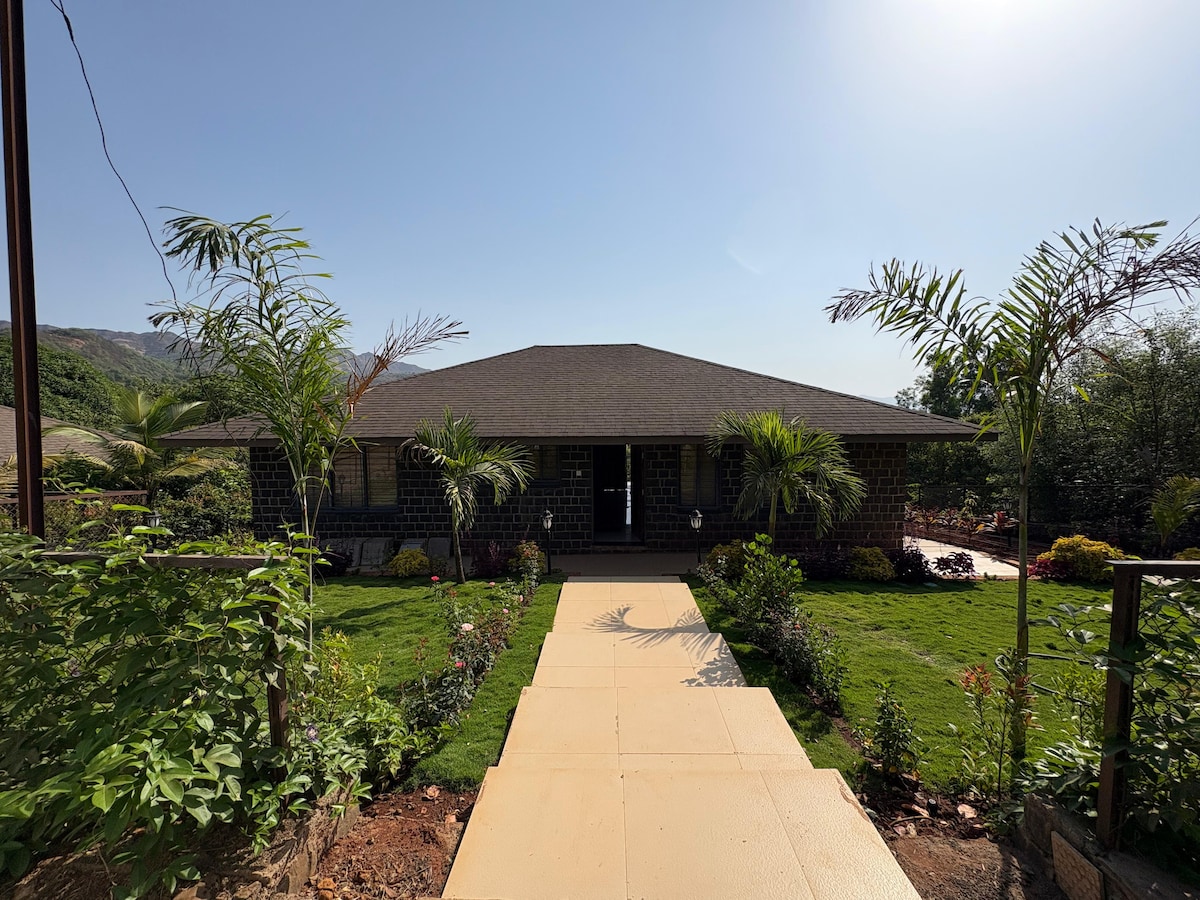 The villa is framed by lush greenery and a well-maintained garden path leading to the entrance. The single-story structure features a covered porch and multiple windows, allowing natural light to fill the space, while palm trees and colorful plants enhance the inviting outdoor setting.