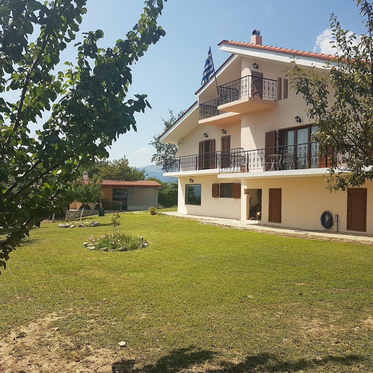 A two-story villa is set amidst a lush green lawn, framed by trees. The exterior features multiple balconies with a Greek flag displayed. Large windows reflect natural light, enhancing the inviting atmosphere of the space. A garden area with minimal landscaping is visible in the foreground.