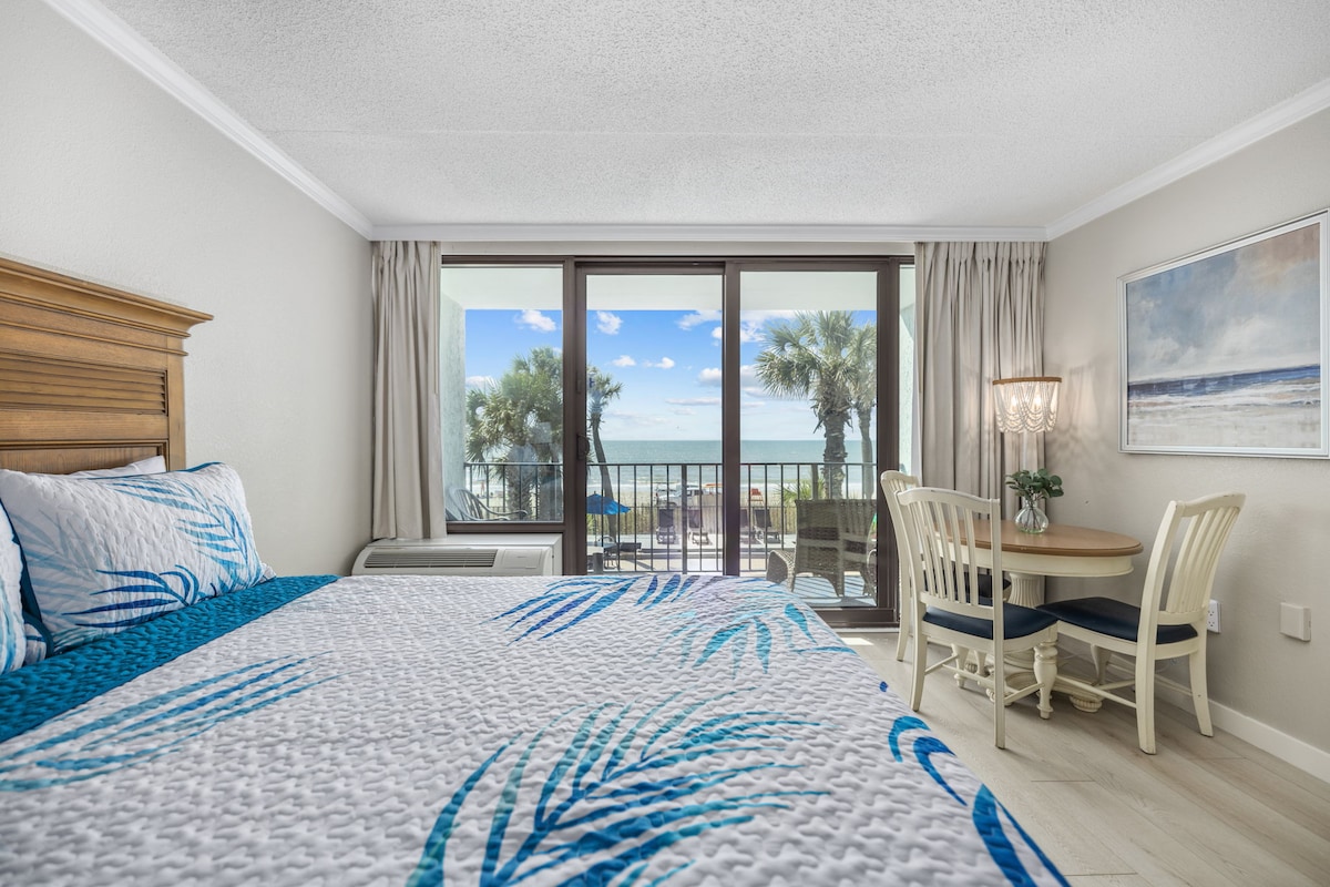 A bright studio features two queen beds adorned with a patterned blue and white quilt. A cozy dining nook is positioned beside the window, offering views of the palm trees and ocean. Sliding glass doors lead to a private balcony, enhancing the airy feel of the space.