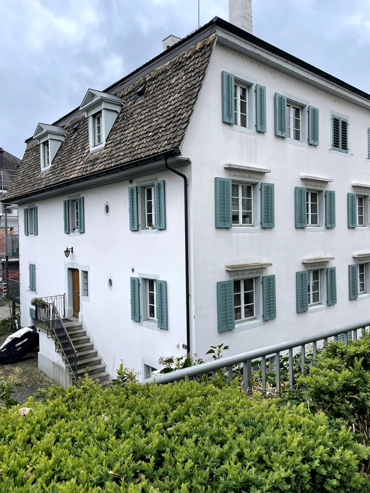 The exterior of a white building with blue shutters is visible, showcasing a charming architectural style. A set of stone steps leads up to the front door, while neatly trimmed greenery surrounds the base of the structure. The roof features a slate design and dormer windows.