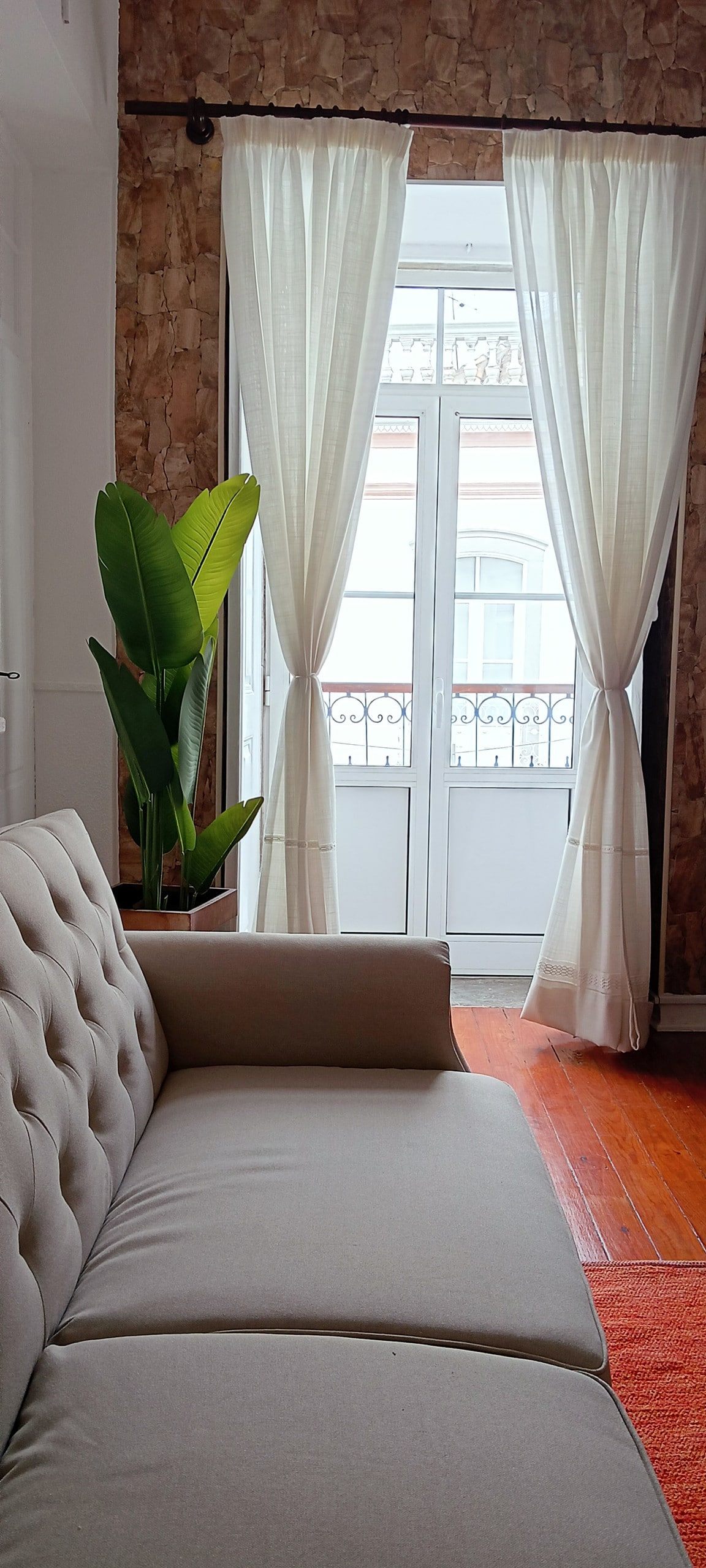 A light-filled room is presented, featuring a comfortable, neutral-toned sofa. Two flowing white curtains frame a glass door that opens to a balcony. A large, green plant is positioned nearby, adding a touch of nature to the inviting space.