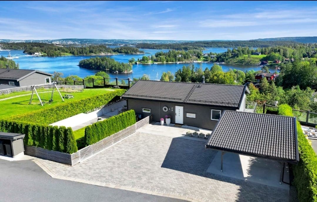 A modern home is situated on a spacious, landscaped property, displaying a sweeping view of the surrounding fjord. The yard features a swing set and well-maintained greenery, while the driveway leads up to the entrance, encapsulated by tall hedges ensuring privacy.