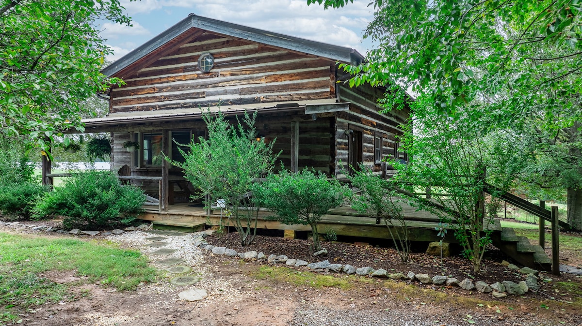 A rustic log cabin is surrounded by lush greenery, offering a welcoming outdoor space. A wooden deck extends from the cabin entrance, while a stone pathway leads through the well-maintained yard, enhancing the natural setting of the property.