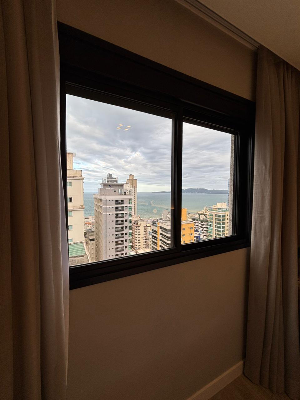 A window framed by neutral-colored curtains provides a view of the cityscape and coastline. Various buildings rise into the sky, while distant water glimmers under a partly cloudy sky, contributing to a sense of urban accessibility.