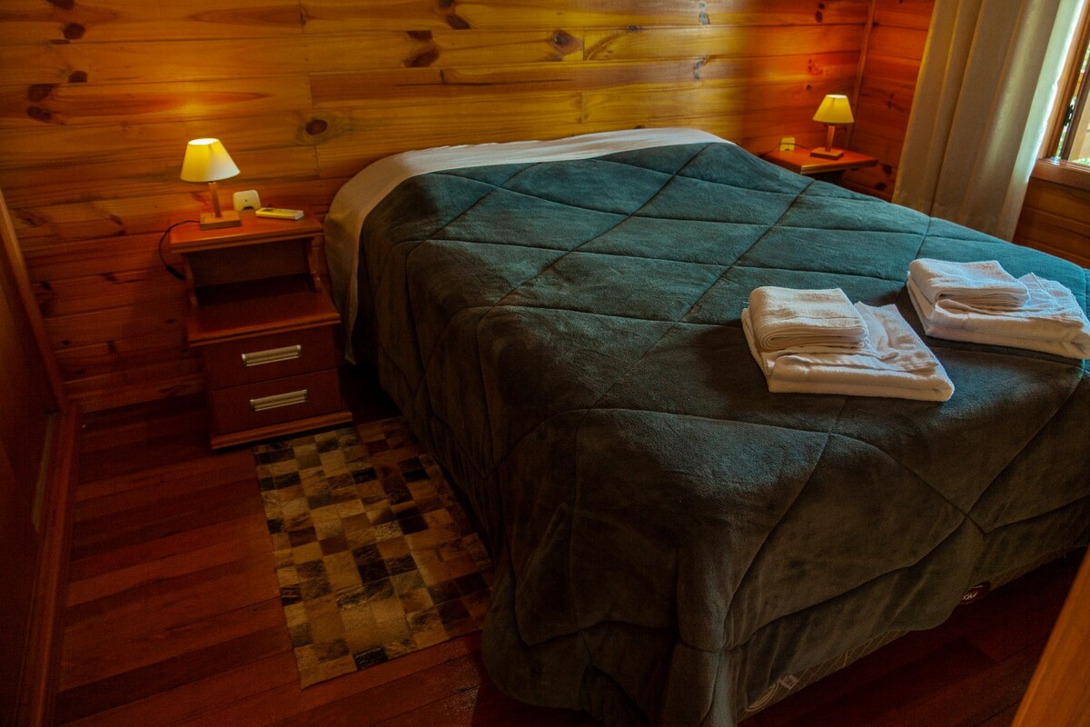 A cozy sleeping area features warm wooden walls and soft lighting. A spacious bed is draped with a textured blanket, complemented by neatly arranged towels. Bedside tables with lamps are positioned on either side, and a small rug adds a touch of character to the wooden floor.