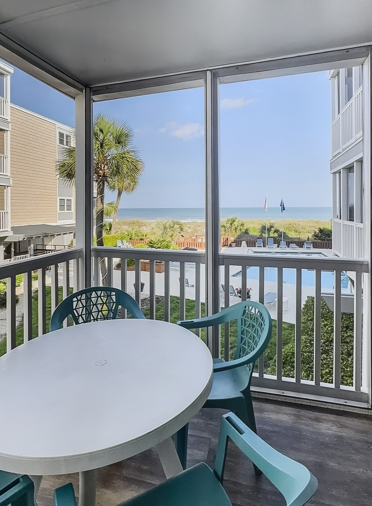 A screened-in porch features a round white table surrounded by four teal chairs, with a view of the ocean visible through the mesh. The sun illuminates the scene, highlighting surrounding palm trees and the pool area below.