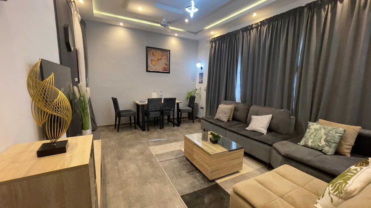 A well-appointed living area features neutral-toned furniture, including a comfortable sectional sofa and a wooden coffee table. Soft lighting from ceiling fixtures provides a cozy ambiance, while large curtains frame the windows. A dining table is set for four, positioned in the background.