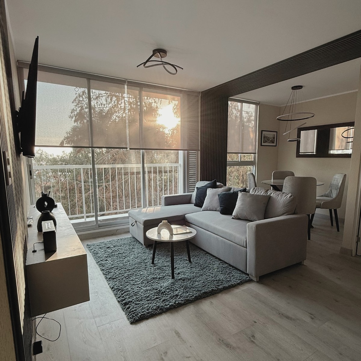 A modern living area is presented, featuring a light grey sectional sofa adorned with decorative pillows. A round coffee table sits on a soft area rug, and large windows allow natural light to fill the space, offering views of the outdoors. A TV is mounted on the wall.