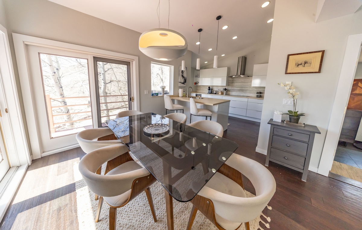 A well-lit dining area features a modern glass table surrounded by six comfortable chairs. Natural light floods through large sliding doors that open to a scenic balcony, while a contemporary light fixture hangs above. The space is complemented by a neutral rug and subtle decorative accents.