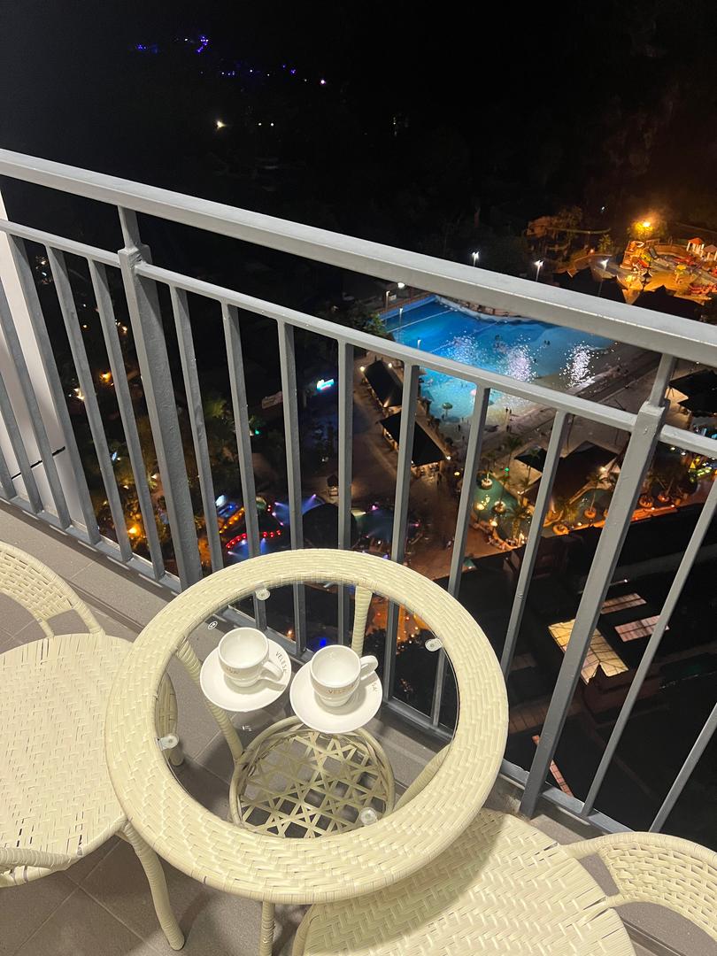 A small outdoor balcony is seen, furnished with two light-colored chairs and a circular table. On the table, two cups are placed. Below, the illuminated resort pool area is visible, featuring colorful lights and various recreational facilities.