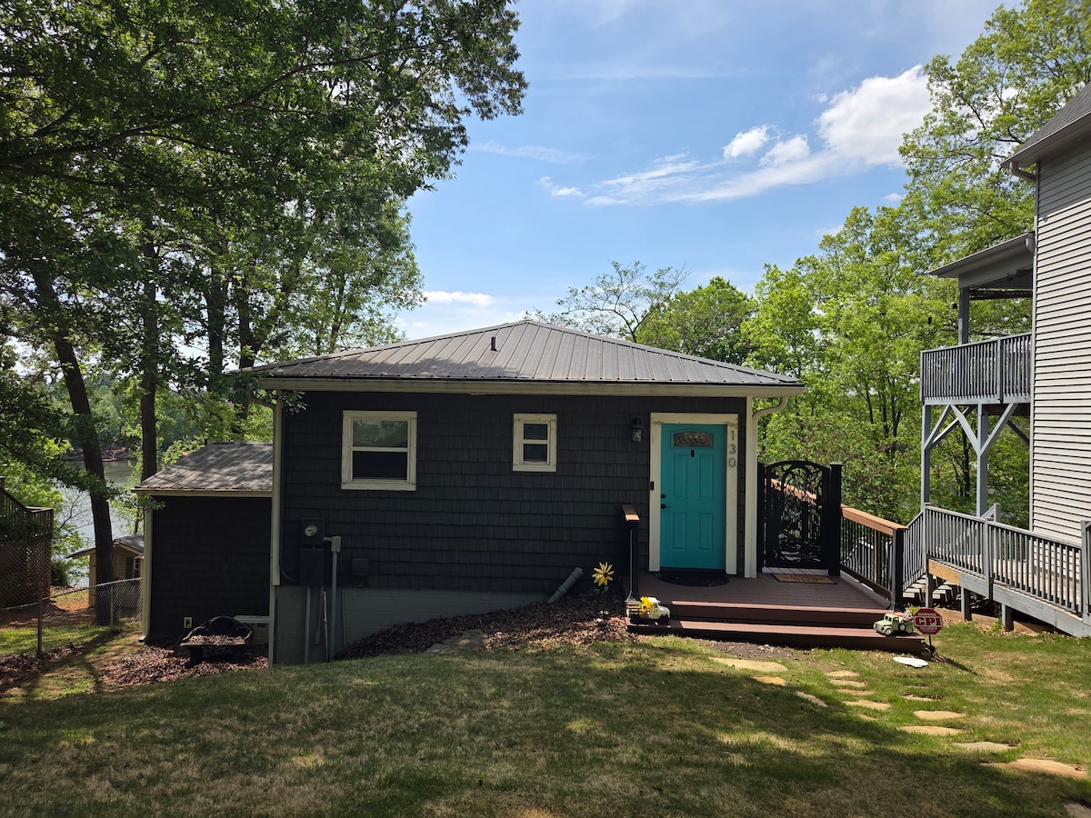 A private home with a vibrant blue door is surrounded by lush greenery, capturing a sense of tranquility. The structure is complemented by a spacious deck, leading down to a pathway framed by natural landscaping, with glimpses of the lake visible in the background.