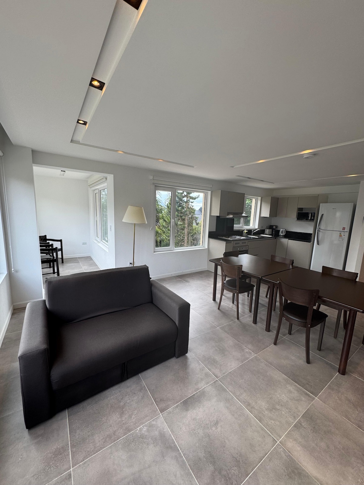 An open living and dining area features a dark sofa adjacent to a wooden dining table with matching chairs. Large windows allow natural light to fill the space, while light fixtures overhead enhance the modern design. A kitchen area is partially visible in the background.