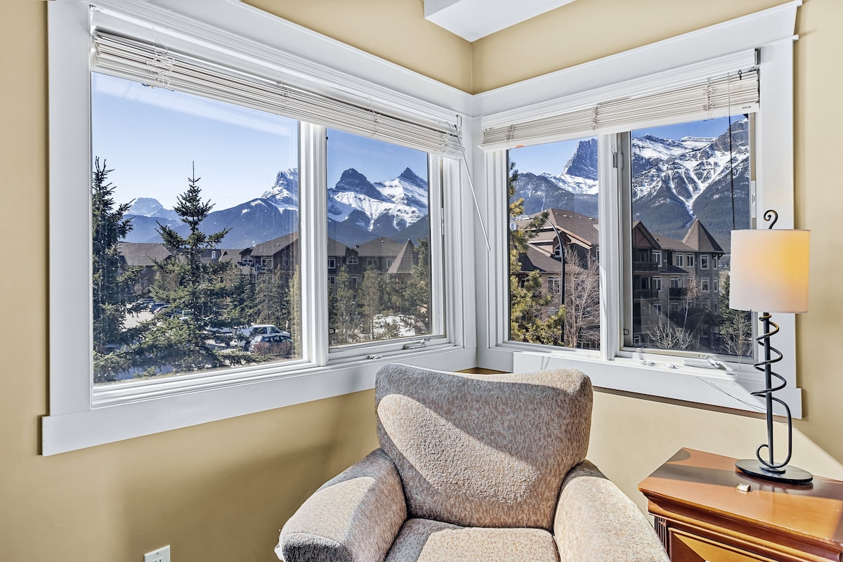 A cozy corner features a softly upholstered accent chair positioned by expansive windows. Natural light floods the space, highlighting mountain views through the glass, while a bedside lamp adds a touch of warmth to the inviting atmosphere.