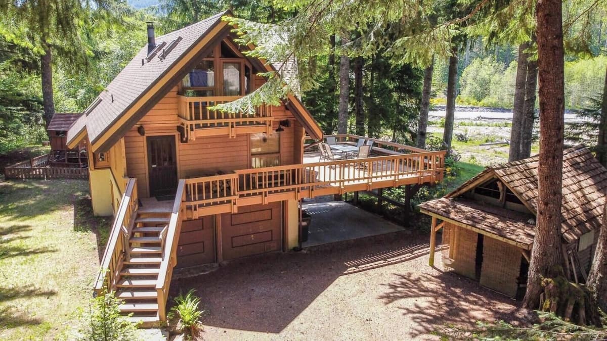 Alpen Commune: a three-level wood chalet with wraparound decks, tall firs and the White River behind it