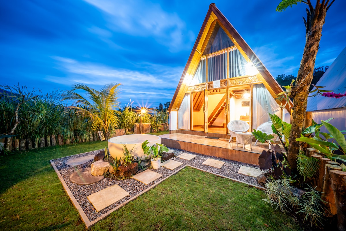 An A-frame structure is set against a dramatic evening sky, featuring large windows that illuminate the interior. A small outdoor seating area is surrounded by landscaped gardens, with stepping stones leading to a circular hot tub, offering a serene environment for relaxation.