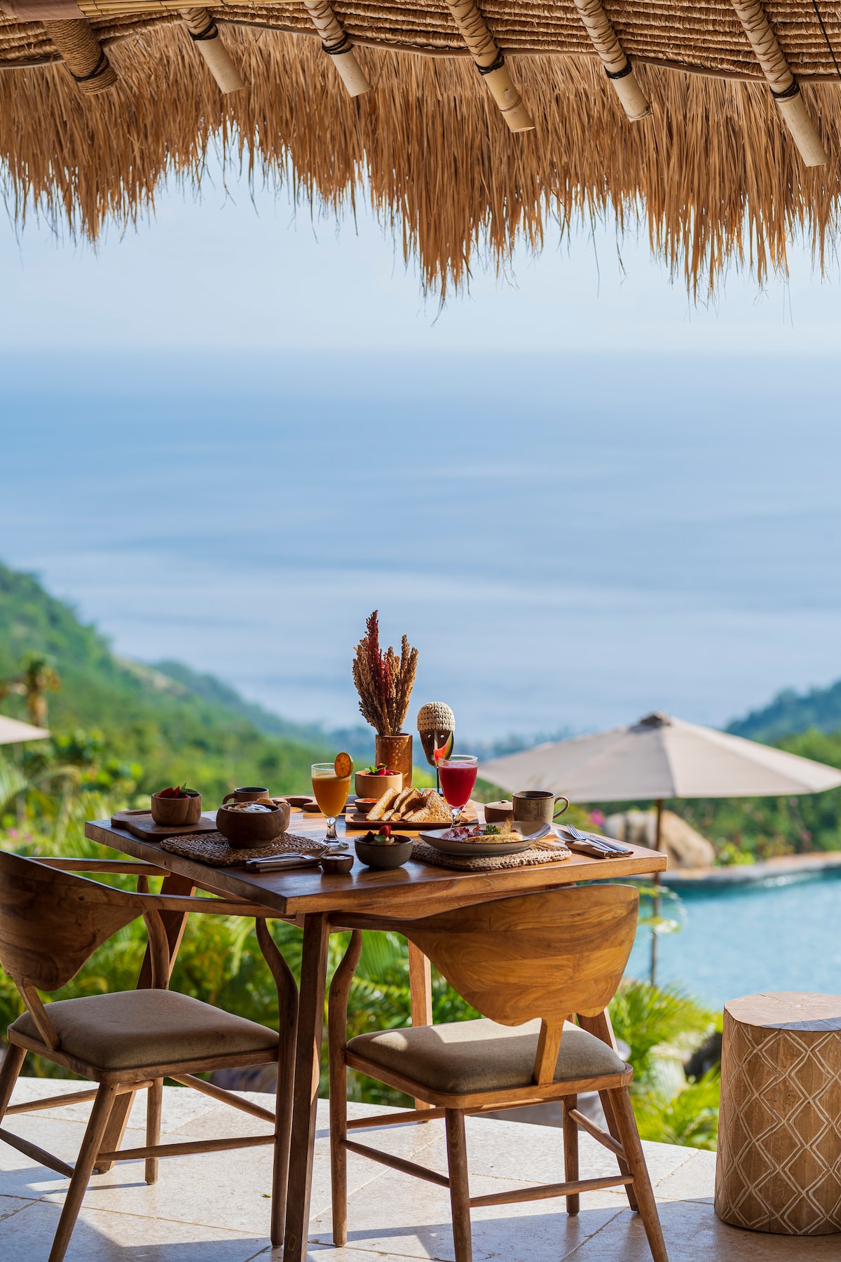 A wooden dining table is set with an array of breakfast items, positioned under a thatched roof. Beyond the table, a tranquil view of the ocean is visible, complemented by a swimming pool and shaded lounge areas in the foreground.