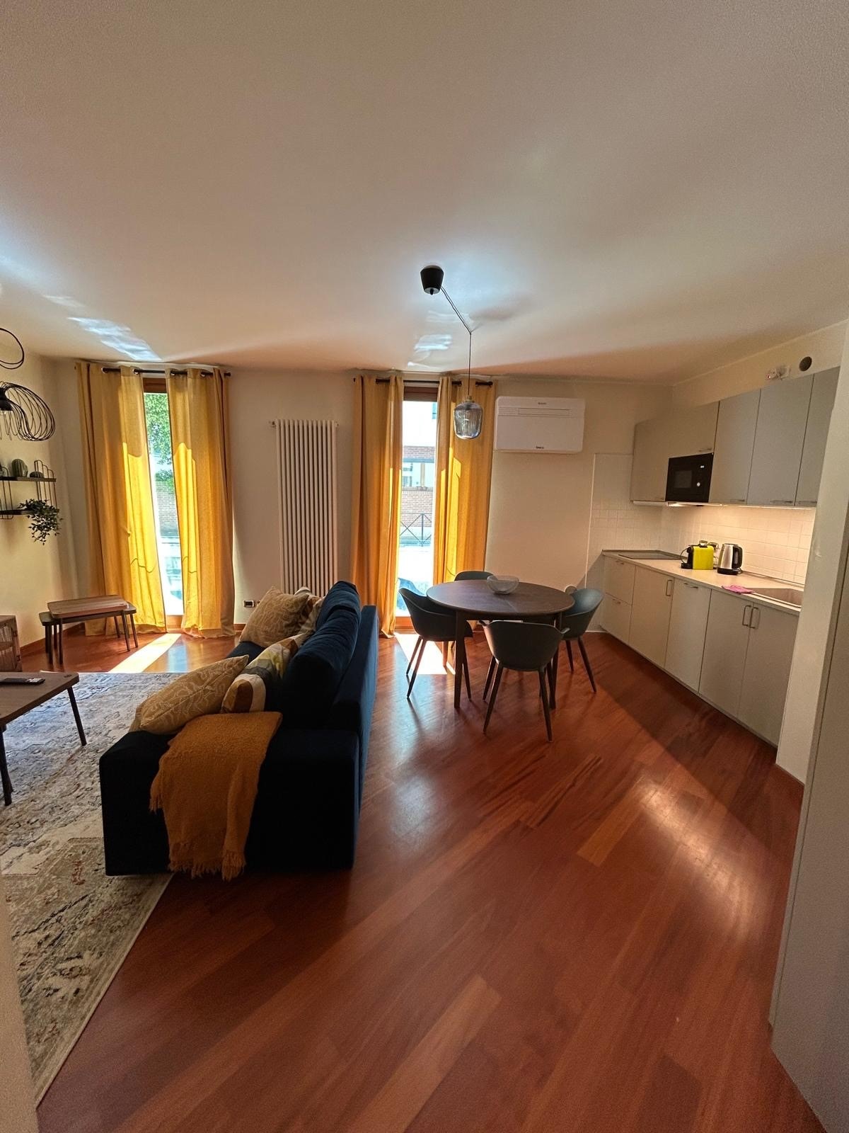 A living area showcases elegant wooden flooring, complemented by a dark blue sofa adorned with various cushions. A round dining table with four chairs is positioned nearby, illuminated by natural light filtering through large windows adorned with yellow curtains. A kitchenette area is visible in the background.