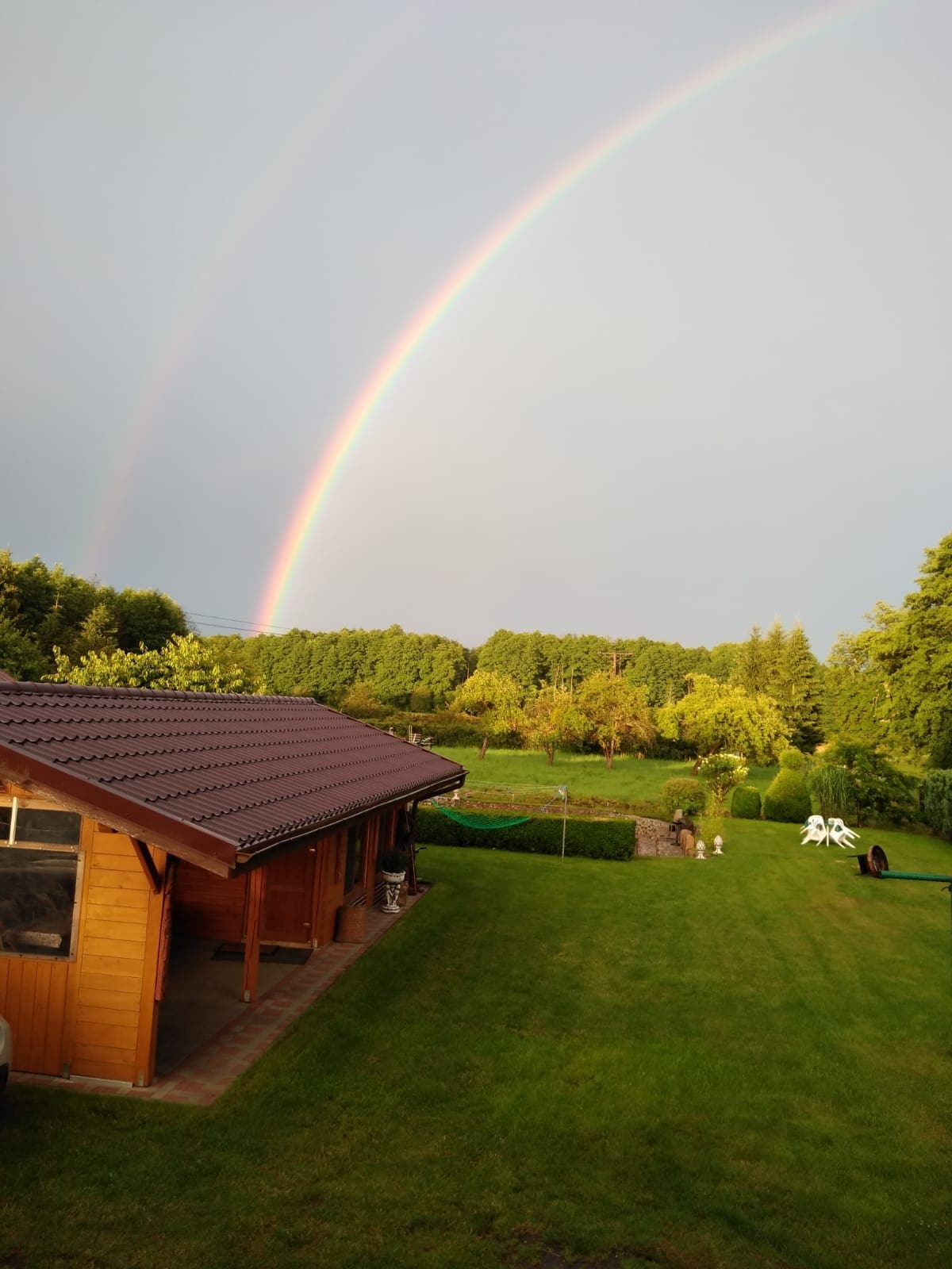 A serene outdoor setting is depicted, with a wooden cabin situated on lush green grass. A double rainbow arcs elegantly over the scene, while trees and garden foliage complement the tranquil landscape.