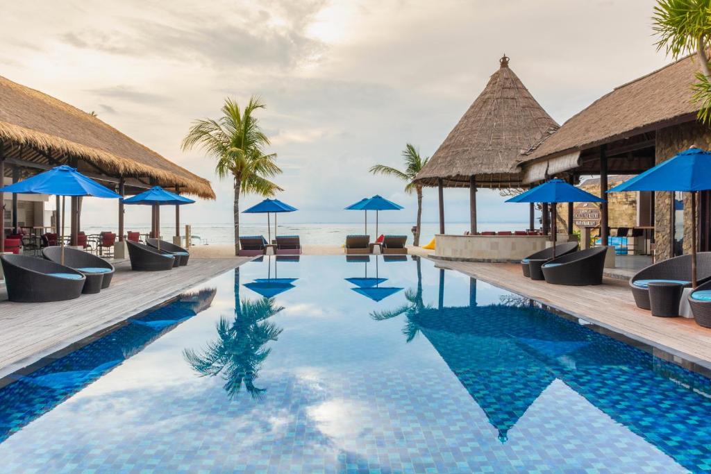 A tranquil outdoor area features a crystal-clear pool, reflecting the surrounding palm trees and thatched-roof structures. Blue umbrellas provide shade over dark lounge chairs arranged near the water's edge, inviting relaxation with ocean views in the background.