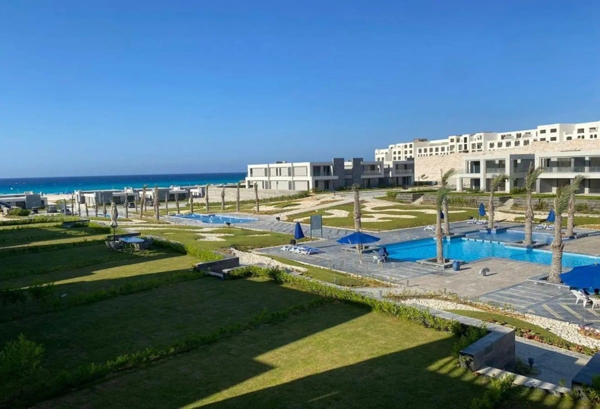 The image captures a serene landscape featuring multiple swimming pools surrounded by lush greenery and sun loungers under blue umbrellas. The view extends to a beach with gentle waves, framed by a clear blue sky, creating an inviting environment for relaxation.