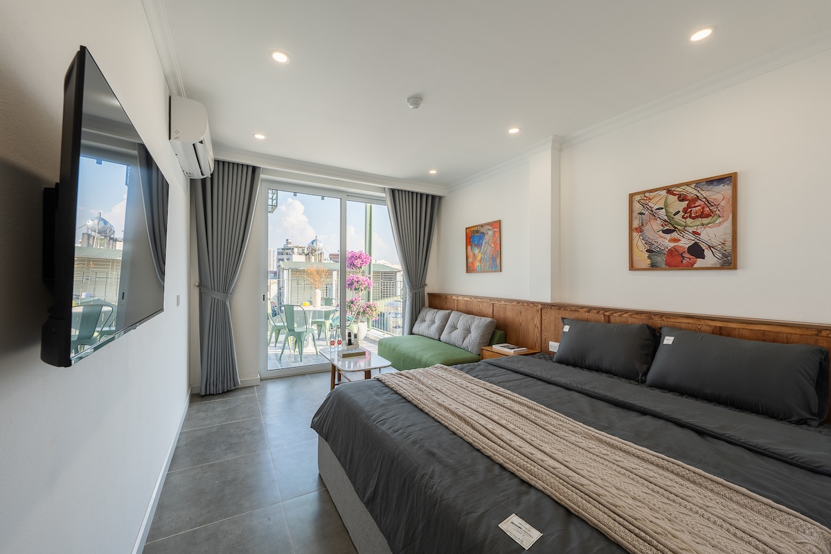 A modern studio apartment features a spacious bed with soft linens, complemented by a cozy seating area. Natural light enters through the large glass doors, which open to a balcony with a scenic view. Vibrant artwork adorns the walls, enhancing the room's contemporary decor.