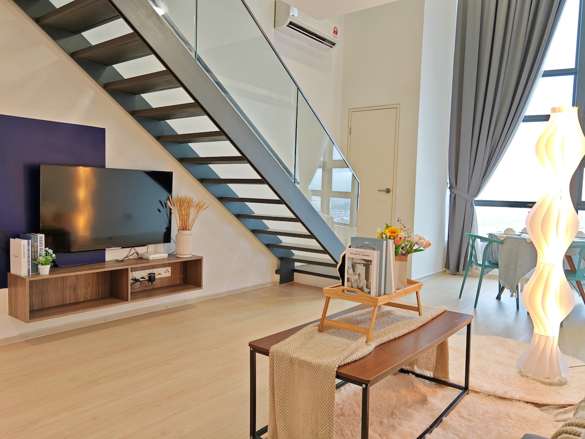 A modern living area features a sleek staircase leading to the upper floor. A flat-screen TV is mounted on the wall above a wooden console. A stylish floor lamp illuminates a coffee table, adorned with a soft throw and a decorative tray holding a small floral arrangement.