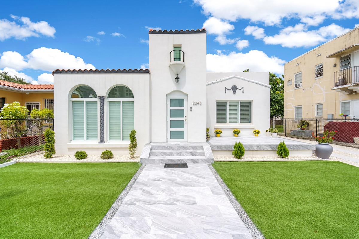 This elegant white home features a unique architectural design with large arched windows and a striking front door. A well-maintained lawn and neatly arranged landscaping enhance the entrance, providing a welcoming approach. The modern exterior is complemented by a contrasting stone pathway leading to the front door.
