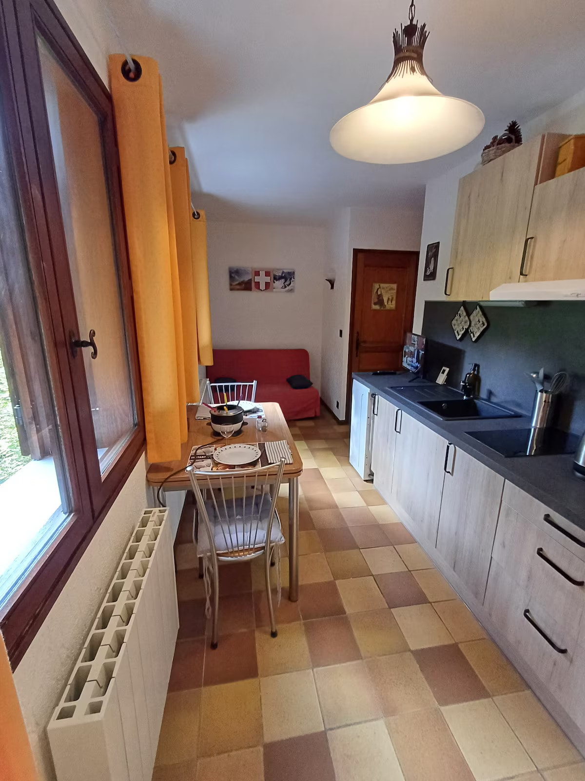 The kitchen area features a compact dining table with two chairs and a folding chair, positioned beside a red couch. Light enters through a window adorned with cheerful yellow curtains. Kitchen cabinets and a black countertop complement the tiled floor, which presents a warm color palette.